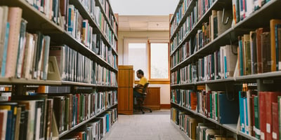 AMBS student sitting in the library at a desk with book shelves on the left and right of the photo