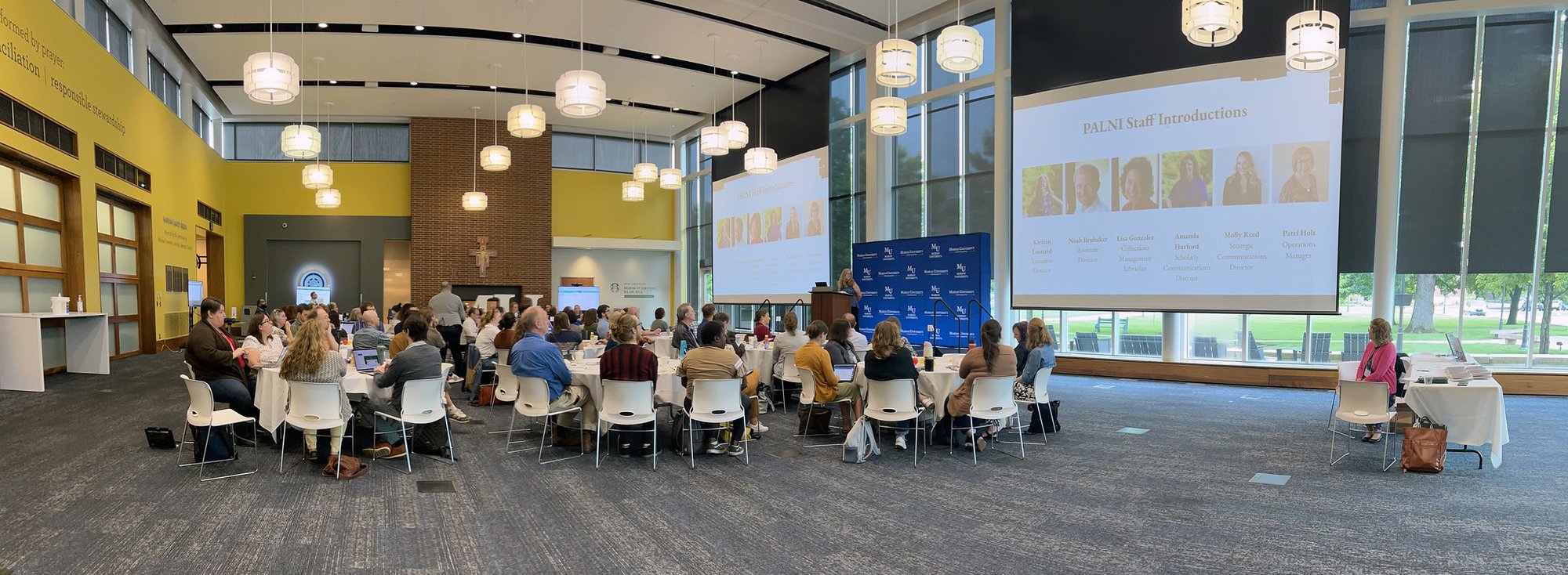 A large group of meeting attendees sitting at tables in a conference room, facing a presenter and two large presentation screens.