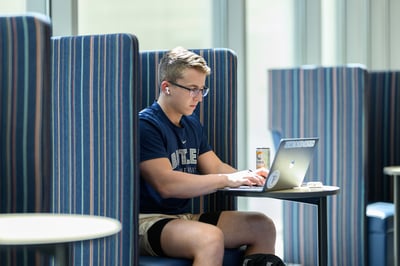 Butler University student sitting at a small table and looking at/typing on a laptop.