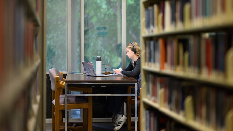 Butler University student sitting at a desk in the library, wearing headphones and looking at an open laptop. The photo is framed on the left and right by two full bookshelves.