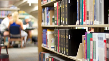 A library aisle with bookshelves filled with books. In the background, people are seated at a table, appearing to study.