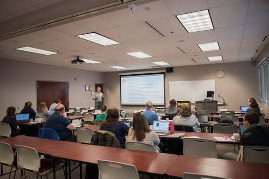 Individuals sitting in a classroom and facing an instructor and projection screen