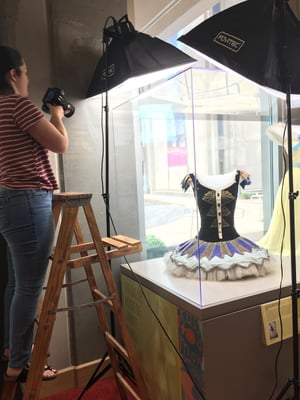 Student standing on a ladder and taking photos of a ballet costume inside a glass box