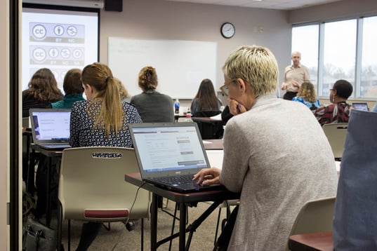 Group of individuals sitting in workshop facing projection screen and looking at laptops as instructor stands at front of room