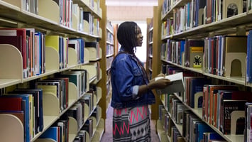 Library patron at Taylor University stands in front of bookshelves and browses titles while holding a book and smiling