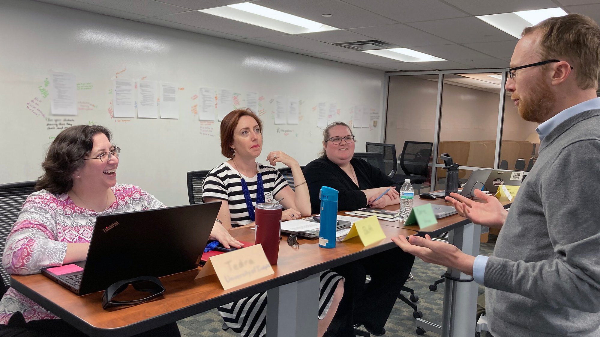 Three individuals sitting at a desk with open laptops and a whiteboard with papers and notes behind them, listening to a person standing and consulting with them.