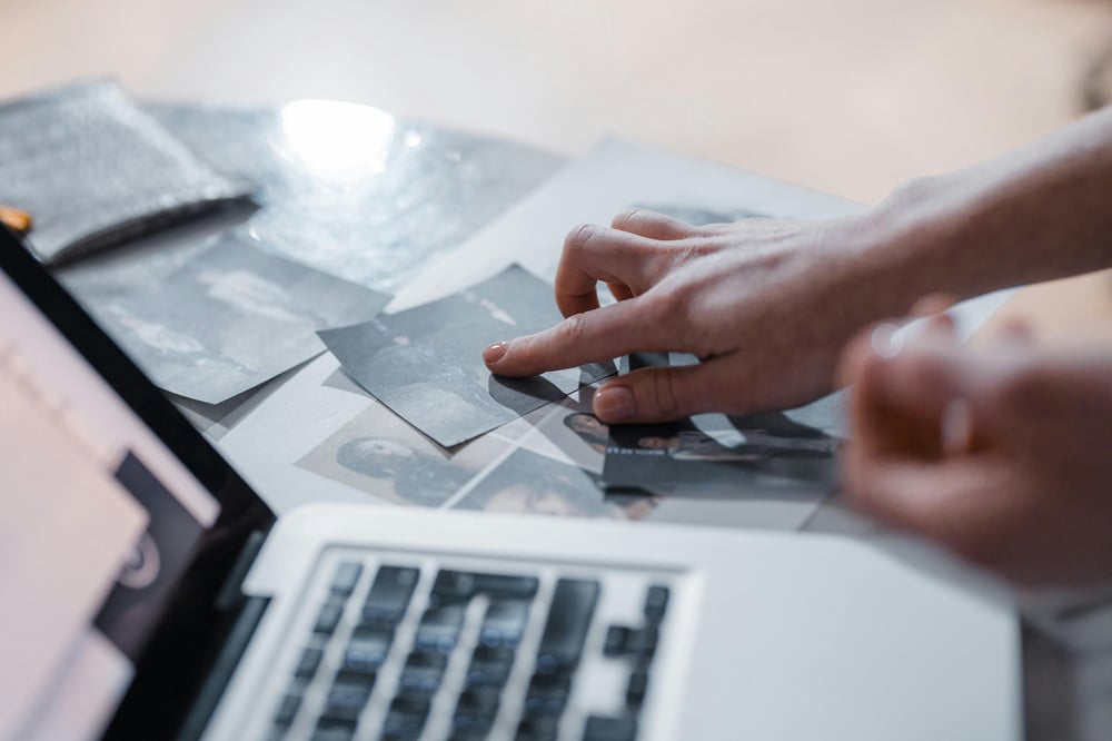 Open laptop next to old photographs with a hand pointing to one of the photos