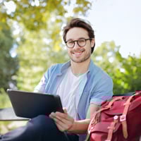 A person sitting outside holding a laptop and smiling with a backpack beside him.