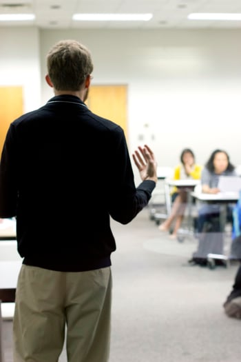 Person with their back facing the camera, standing in front of a classroom and appearing to be instructing students.