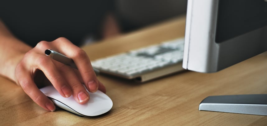 person's hand holding a pen and moving a mouse next to a keyboard in front of a computer monitor on a desk.