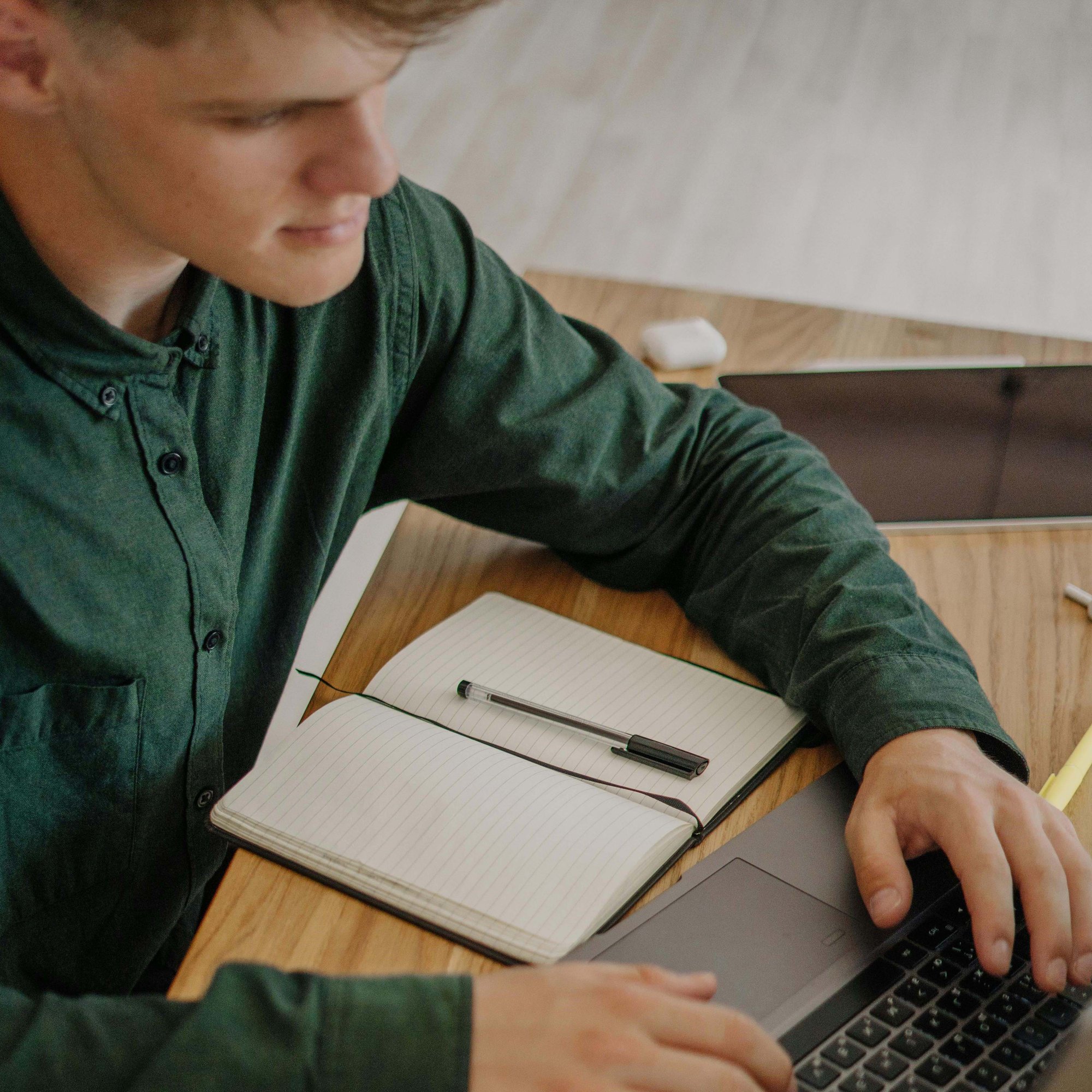 College-age student working on a laptop with an open notebook in front of them