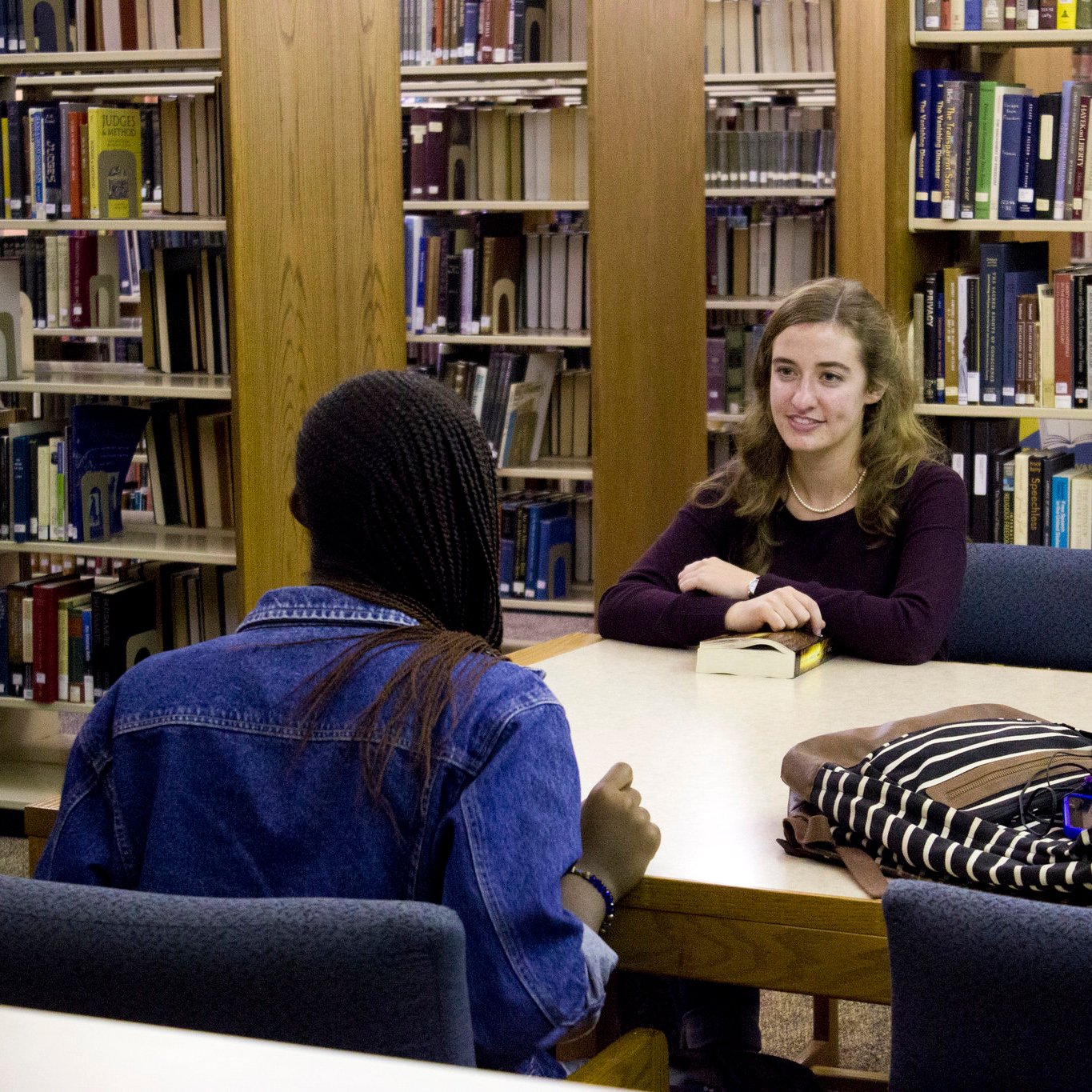 Two college students sitting at a desk inside a library, having a conversation.