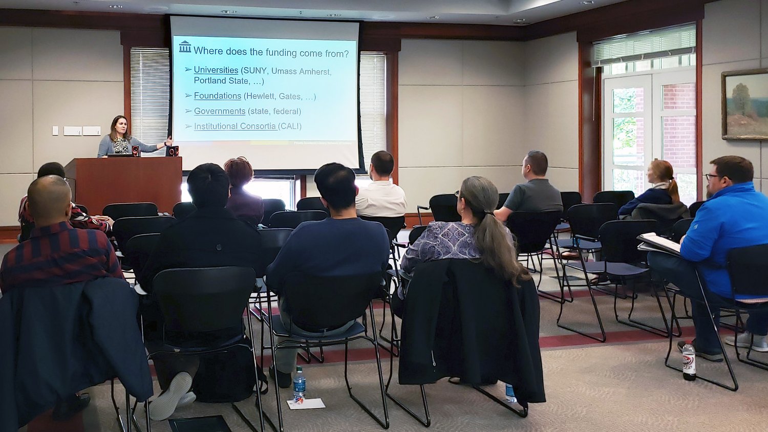 An instructor stands behind a podium and points to a white board, addressing several adult learners sitting at desks.